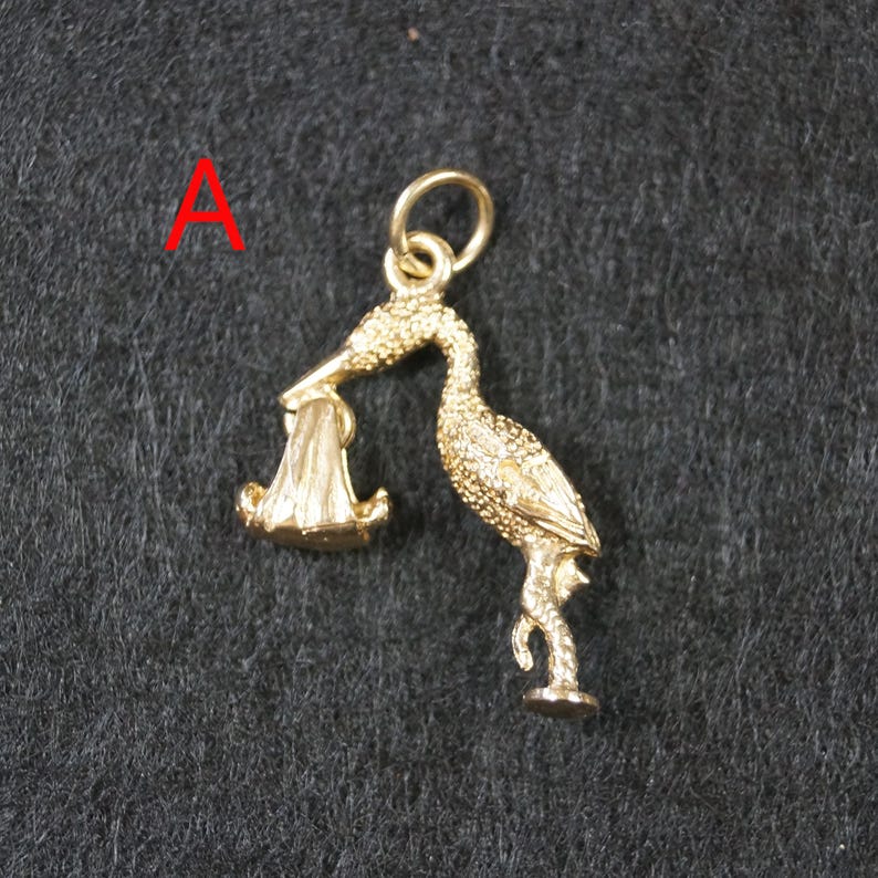 May include: A gold-colored metal charm in the shape of a stork carrying a bundle in its beak. The stork is depicted standing on one leg with textured feather details across its body. A small circular jump ring is attached to the top of the stork.