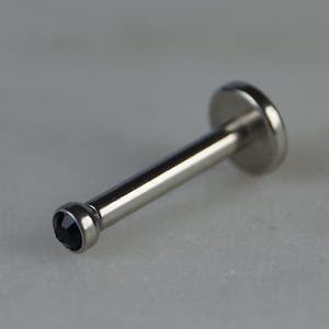 Könnte beinhalten: Ein silberfarbener Lippenpiercing-Stecker mit einer flachen Scheibe an einem Ende und einem kleinen, runden, schwarzen Edelstein am anderen. Das Piercing ist für die Lippe konzipiert und besteht aus glänzendem Metall.