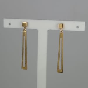 May include: A pair of gold-toned drop earrings featuring a small square stud connected to a long, slender, trapezoidal frame with an open center. The earrings have a smooth, polished finish and hang vertically from a white display stand.