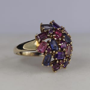 Op de afbeelding: Een gouden ring met een cluster van kleurrijke edelstenen. De ring heeft rechthoekige en ronde stenen in roze, paarse en blauwe tinten. De ring heeft een eenvoudige, gepolijste gouden band.