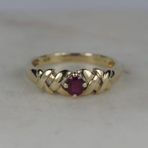Può includere: Un anello in oro con un rubino rosso centrale a taglio rotondo. La fascia presenta una finitura in oro lucido e un motivo decorativo incrociato su entrambi i lati della pietra, che è fissata da una montatura a quattro griffe.