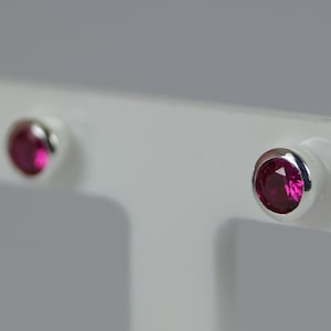 May include: A pair of stud earrings with bright magenta gemstones set in a polished silver bezel. The earrings are displayed against a white background, highlighting their simple elegance and vibrant colour.