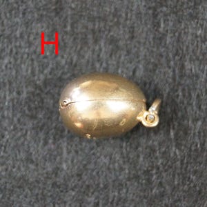 May include: A small, oval-shaped gold-colored pendant with a smooth, metallic finish and a hinged seam running horizontally across the center. A small circular bail is attached to one side for hanging. The letter H is visible in red text.