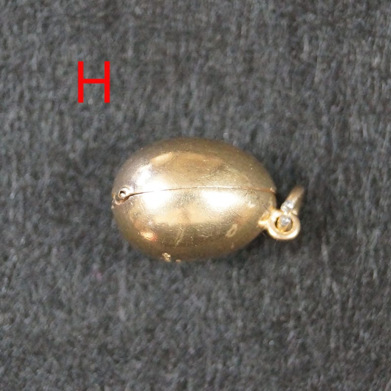 May include: A small, oval-shaped gold-colored pendant with a smooth, metallic finish and a hinged seam running horizontally across the center. A small circular bail is attached to one side for hanging. The letter H is visible in red text.