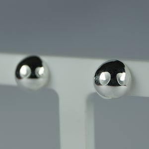 May include: A pair of polished silver stud earrings. Each earring is a smooth, rounded dome shape, reflecting the light. The earrings are displayed on a white surface against a grey background. Simple and classic jewellery.