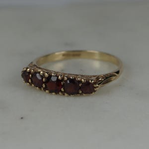 May include: A gold ring featuring five round, deep red garnet gemstones set in a row. The band has an intricate, decorative filigree pattern along the sides, and the stones are held in place by small, rounded metal prongs in a classic setting.