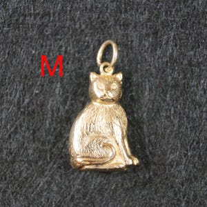 May include: A gold-colored metal pendant shaped like a sitting cat with textured fur details and a small circular jump ring at the top for attachment. The letter M is printed in red to the left of the cat pendant, which is centered in the frame.