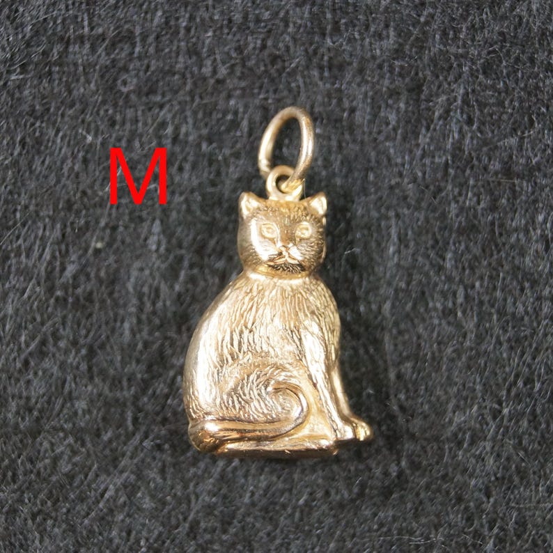 May include: A gold-colored metal pendant shaped like a sitting cat with textured fur details and a small circular jump ring at the top for attachment. The letter M is printed in red to the left of the cat pendant, which is centered in the frame.