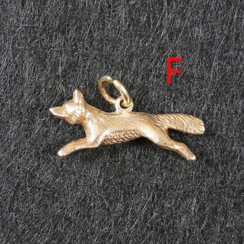 May include: A gold-toned metal charm shaped like a running fox with textured fur details and a small circular jump ring attached to the top. The letter F is printed in red in the upper right corner of the frame, positioned above the fox charm.