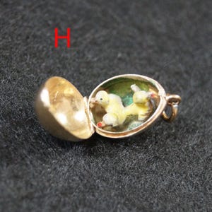May include: A gold-toned metal charm shaped like an egg that opens to reveal a small, yellow, painted duckling figure inside. The charm features a hinged lid and a small loop at the side for attachment. The letter H is visible in red text.