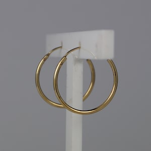 May include: A pair of thin, circular gold hoop earrings with a smooth, polished finish. The earrings are suspended from a white display stand, showcasing their simple, round design and metallic yellow colour in a clear, focused view.
