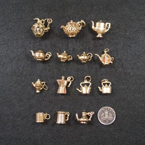 May include: A collection of fourteen gold-toned metal charms shaped like teapots, kettles, and mugs, arranged in four rows. A circular British five-pence coin is included for scale, showing the text 5 and a crown design on its metallic surface.