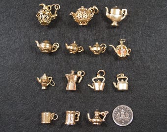May include: A collection of fourteen gold-toned metal charms shaped like teapots, kettles, and mugs, arranged in four rows. A circular British five-pence coin is included for scale, showing the text 5 and a crown design on its metallic surface.