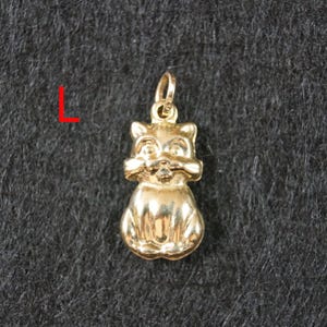 May include: A gold-toned metal pendant shaped like a seated cat with pointed ears and a small bail at the top for attachment. The letter L is printed in red to the left of the pendant, which features a smooth, reflective surface finish.