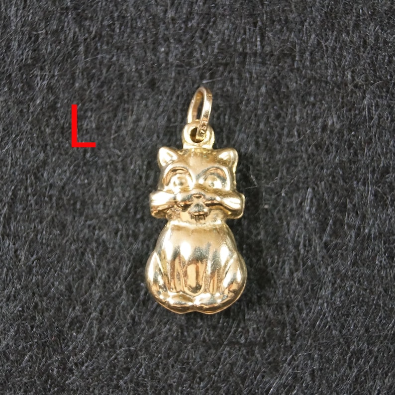 May include: A gold-toned metal pendant shaped like a seated cat with pointed ears and a small bail at the top for attachment. The letter L is printed in red to the left of the pendant, which features a smooth, reflective surface finish.