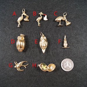 May include: A collection of gold-toned metal charms labeled A through H, including three stork figures, three owl figures, a bird in flight, and an open locket containing a small figure, displayed alongside a circular silver-colored coin.