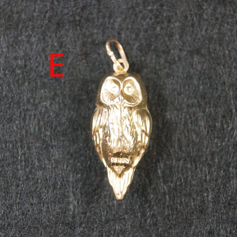 May include: A gold-colored metal owl pendant featuring detailed feather textures and a small circular bail at the top for attachment. The letter E is printed in red to the left of the pendant, which has a polished finish and a distinct beak.