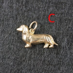 May include: A gold-toned metal charm shaped like a dachshund dog in a standing position, featuring a textured surface to mimic fur and a small circular jump ring attached to the top. The letter C is printed in red in the upper right corner.