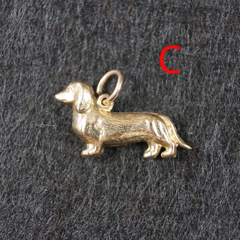 May include: A gold-toned metal charm shaped like a dachshund dog in a standing position, featuring a textured surface to mimic fur and a small circular jump ring attached to the top. The letter C is printed in red in the upper right corner.