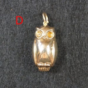 May include: A gold-toned metal owl pendant featuring a small jump ring at the top for attachment. The owl has two faceted yellow stones set as eyes and a smooth, polished body. The letter D is printed in red to the left of the owl pendant.
