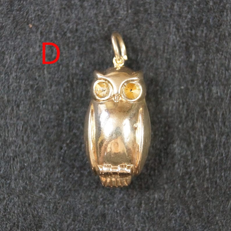 May include: A gold-toned metal owl pendant featuring a small jump ring at the top for attachment. The owl has two faceted yellow stones set as eyes and a smooth, polished body. The letter D is printed in red to the left of the owl pendant.
