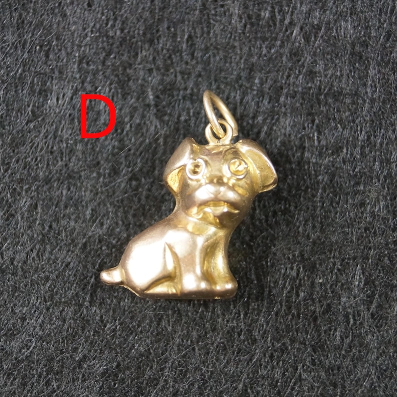 May include: A gold-toned metal charm in the shape of a seated dog with floppy ears and a small jump ring at the top for attachment. The letter D is printed in red to the left of the dog charm, which features a smooth, reflective surface.