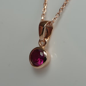 Può includere: Una collana in oro rosa con un pendente rotondo con pietra preziosa rosso intenso. Il pendente è incastonato in una lunetta e pende da una delicata catena. Il gioiello è esposto su sfondo bianco.