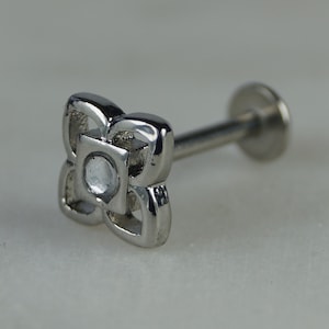 Puede incluir: Un piercing labial plateado con un diseño de flor de cuatro pétalos. El centro de la flor tiene una pequeña gema redonda y transparente. El piercing tiene una parte posterior plana y un poste recto.