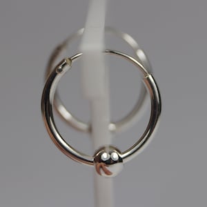 May include: A pair of small, shiny silver hoop earrings. One earring features a small, round bead at the bottom. The earrings are displayed against a light gray background, highlighting their polished surface and simple design.