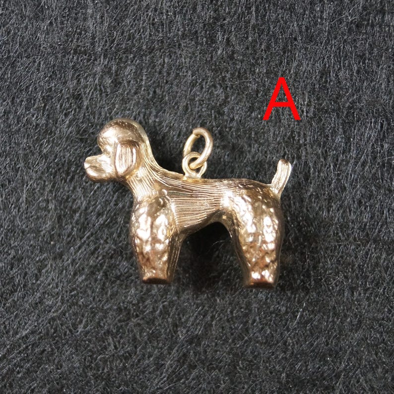 May include: A gold-toned metal charm in the shape of a standing poodle dog, featuring textured fur detailing on the body and a small circular jump ring attached to the back. The letter A is printed in red in the upper right corner of the image.