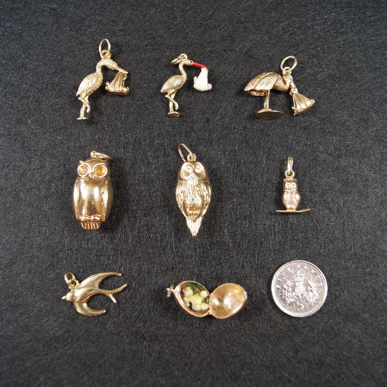 May include: A collection of gold-toned charms including three storks, three owls of varying sizes, a bird in flight, and an open locket containing a small figure, displayed alongside a silver-colored coin for scale on a dark textured surface.