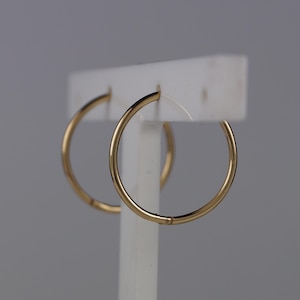 May include: A pair of gold hoop earrings with a smooth, polished finish. The circular earrings are thin and feature a simple, continuous design. They are displayed on a white stand, showcasing their reflective metallic surface and circular shape.