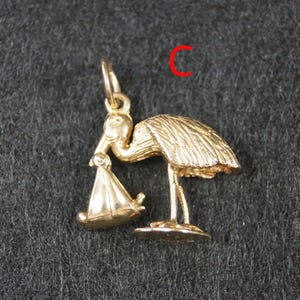 May include: A gold-toned metal charm in the shape of a stork carrying a bundle in its beak. The bird is depicted standing on a small base with detailed feather textures. A small jump ring is attached to the top. The letter C is visible in red.