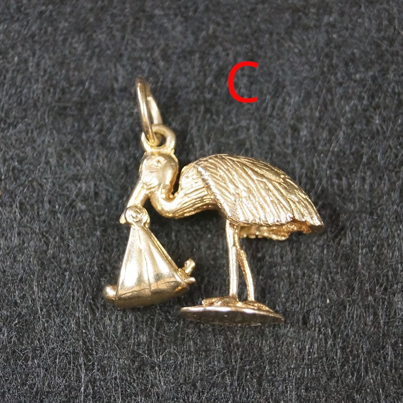 May include: A gold-toned metal charm in the shape of a stork carrying a bundle in its beak. The bird is depicted standing on a small base with detailed feather textures. A small jump ring is attached to the top. The letter C is visible in red.