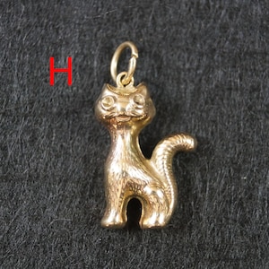 May include: A gold-toned metal cat charm featuring a standing feline figure with a curved tail and a small circular jump ring attached to the top of its head. The letter H is printed in red to the left of the cat charm.