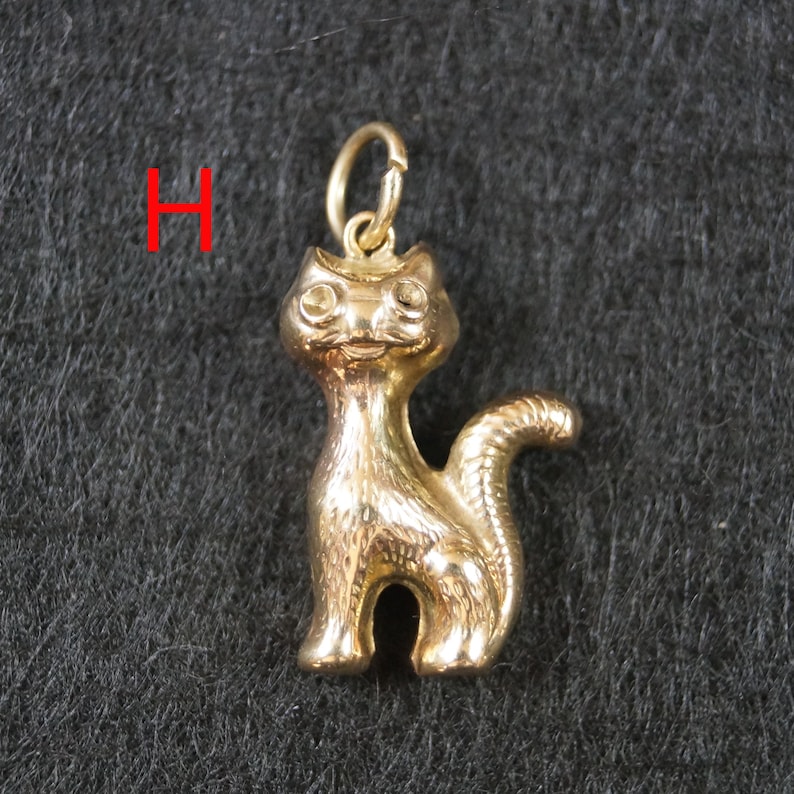 May include: A gold-toned metal cat charm featuring a standing feline figure with a curved tail and a small circular jump ring attached to the top of its head. The letter H is printed in red to the left of the cat charm.