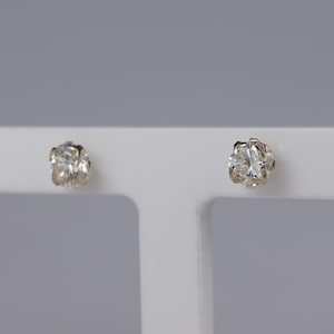 May include: Two clear, round-cut diamond stud earrings. The diamonds are set in a silver or white metal setting. The earrings are displayed on a white surface against a gray background. The earrings are a classic jewelry item.