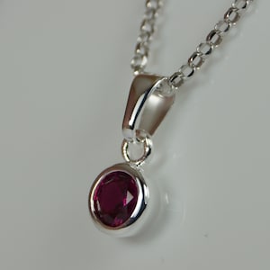 May include: A silver pendant necklace with a round, deep red gemstone. The gemstone is set in a polished silver bezel, suspended from a delicate silver chain. The pendant's design is simple and elegant, suitable for everyday wear.