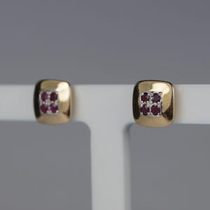 May include: A pair of gold-coloured square stud earrings. Each earring has a cluster of small, deep red gemstones set within a square frame. The earrings are displayed on a white stand, highlighting their design and colour.