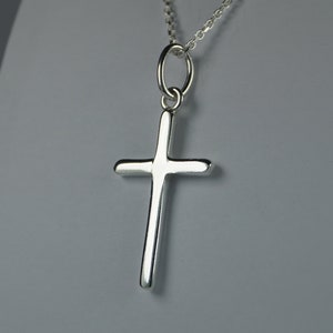 Può includere: Un pendente a forma di croce in argento su una catenina sottile. La croce ha un design semplice e lucido con bordi arrotondati. La collana è mostrata su uno sfondo grigio.