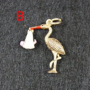 May include: A gold-toned metal charm shaped like a stork carrying a white bundle with a small pink figure inside. The stork has a bright red beak and a textured body. The letter B is printed in red in the upper left corner of the image.