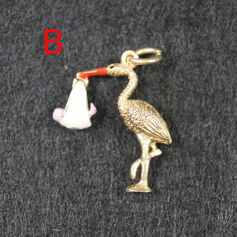 May include: A gold-toned metal charm shaped like a stork carrying a white bundle with a small pink figure inside. The stork has a bright red beak and a textured body. The letter B is printed in red in the upper left corner of the image.