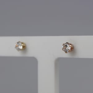 May include: Two stud earrings. One earring has a gold setting with a clear, round gemstone. The other earring has a rose gold setting with a clear, round gemstone. Both earrings are displayed on a white surface.