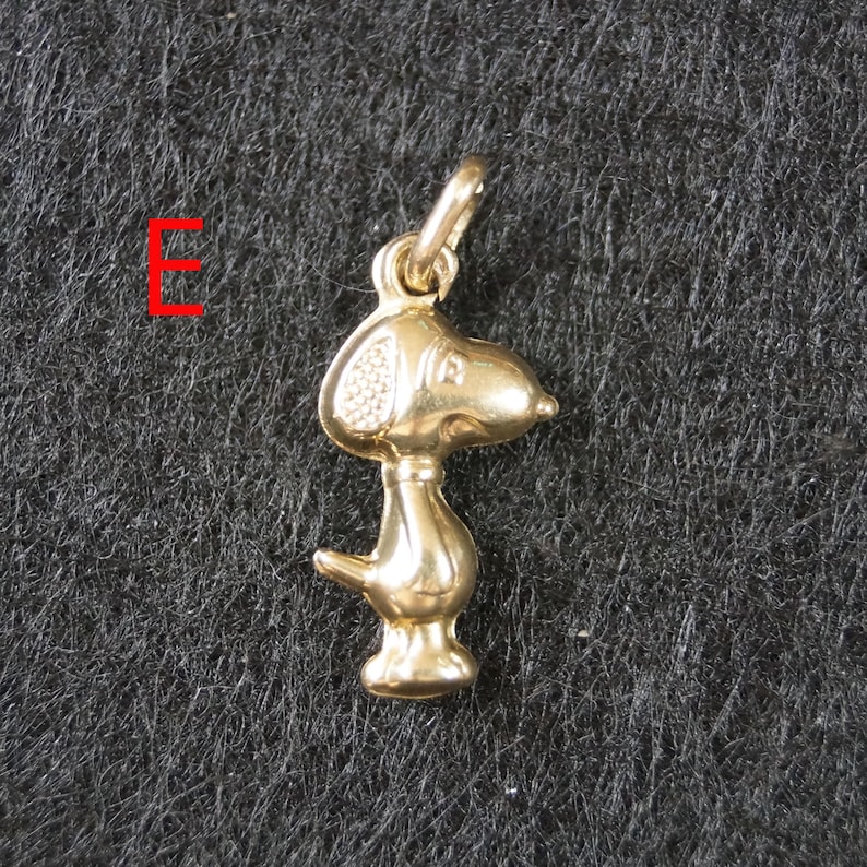 May include: A gold-toned metal pendant shaped like a cartoon dog with a large head, floppy ear, and small tail. The pendant features a small jump ring at the top for attachment. The letter E is printed in red to the left of the dog-shaped charm.