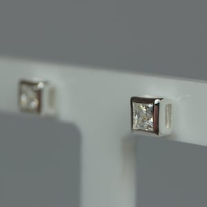 May include: Square-cut diamond stud earrings. The earrings feature a square, clear gemstone set in a silver-coloured metal. The earrings are displayed on a white stand against a grey background.