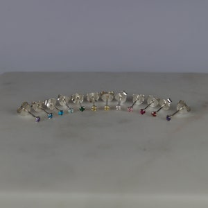May include: A collection of stud earrings with various colored gemstones. The earrings are arranged in a row, each with a different colored gemstone, including purple, blue, green, and pink. The earrings are made of silver.
