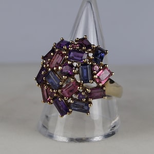 Può includere: Un anello d'oro con un gruppo di pietre preziose colorate. Il design dell'anello include pietre rettangolari e rotonde nelle tonalità del viola, rosa e blu. L'anello è esposto su un supporto conico trasparente.