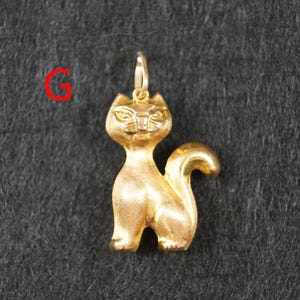 May include: A gold-colored cat pendant featuring a matte finish, pointed ears, and a curved tail. The pendant includes a small jump ring at the top for attachment. The letter G is printed in red in the upper left corner of the image.