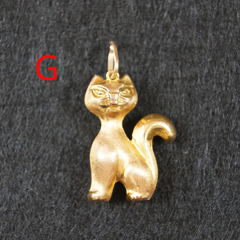 May include: A gold-colored cat pendant featuring a matte finish, pointed ears, and a curved tail. The pendant includes a small jump ring at the top for attachment. The letter G is printed in red in the upper left corner of the image.