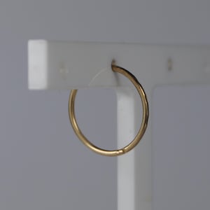 May include: A thin, circular gold hoop earring with a smooth, polished finish. The earring is suspended from a white display stand, showcasing its simple, continuous metal band design and reflective surface quality.